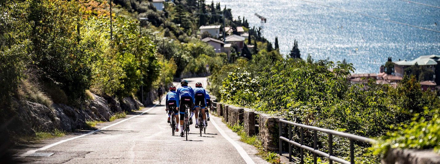 Tre ciclisti su una strada accanto a un lago con montagne sullo sfondo