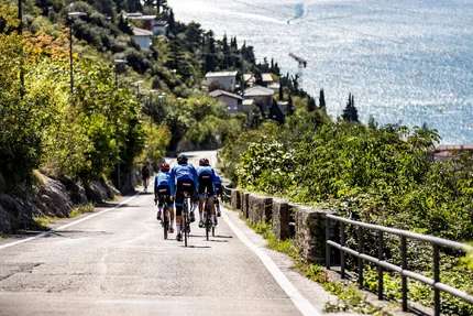 Tre ciclisti su una strada accanto a un lago con montagne sullo sfondo