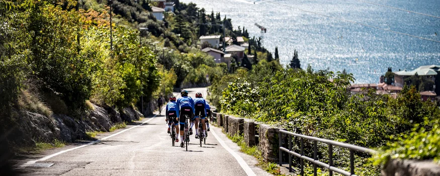 Tre ciclisti su una strada accanto a un lago con montagne sullo sfondo