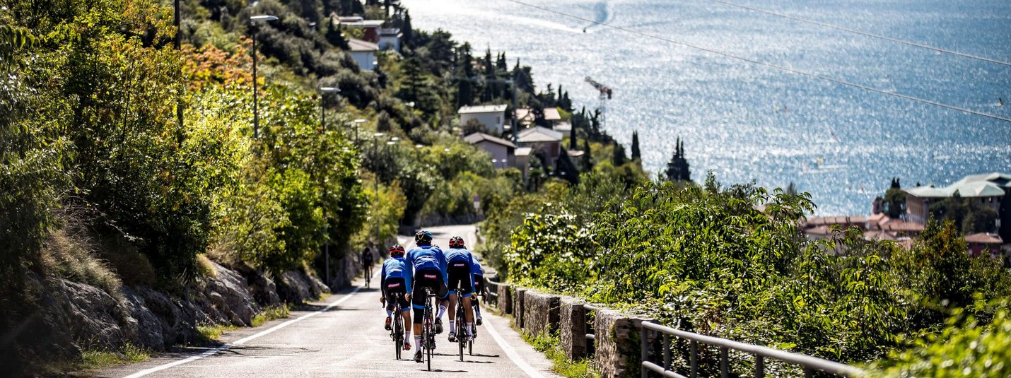 Drie fietsers op een weg langs een meer met bergen op de achtergrond