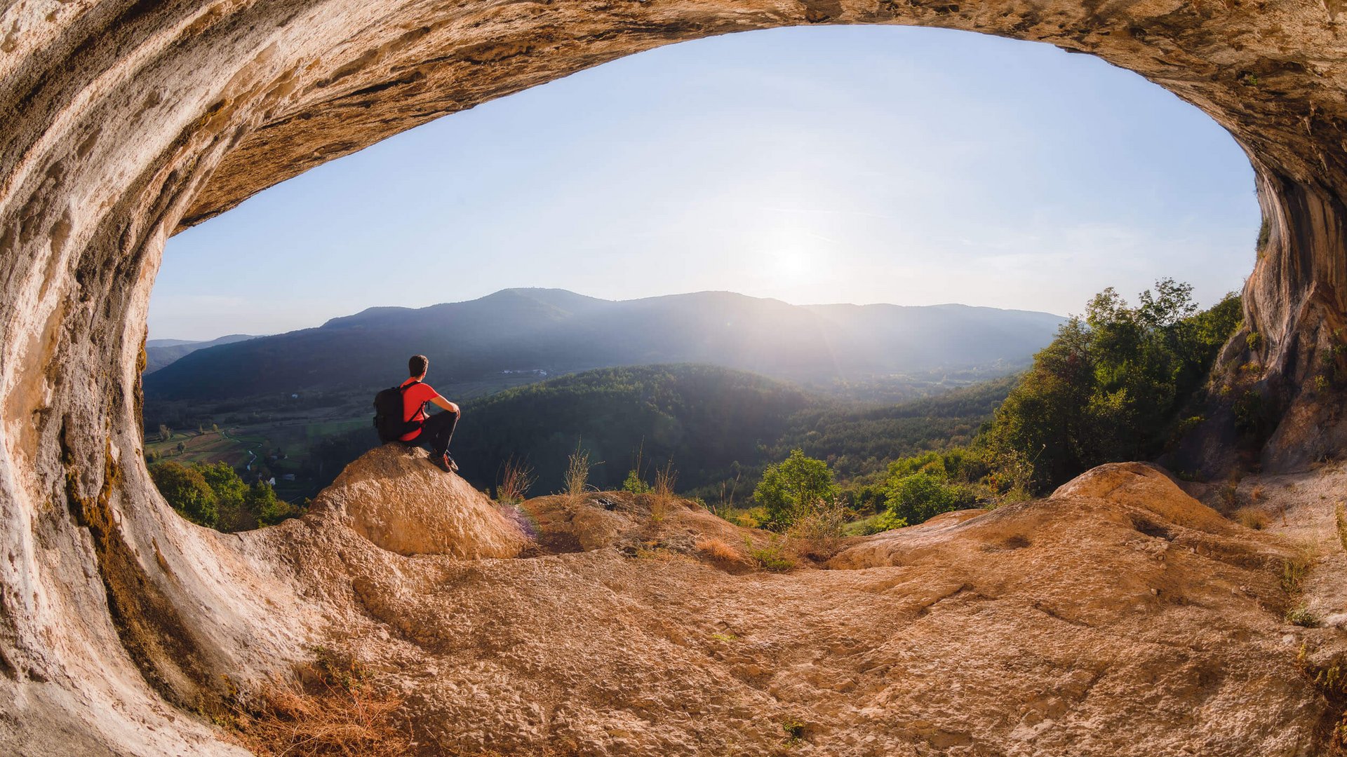 Person sitzt in einer Höhle und blickt auf sonnige Berglandschaft
