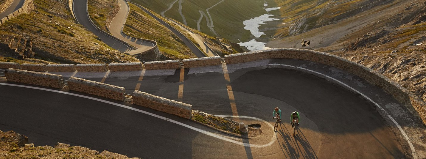 Two cyclists riding a winding mountain road in sunlight