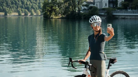 Die schönsten Rennrad-Regionen und Tipps für den Herbst © Alain Rumpf Frau mit Fahrrad macht Selfie am See mit Berg und Schloss im Hintergrund