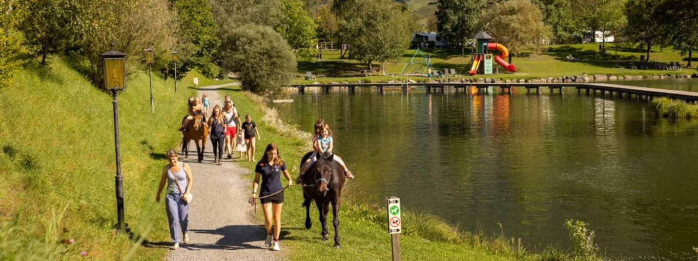 People horseback riding and walking along a lakeside path in a green landscape