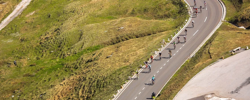 Cyclists on winding mountain road with green grass and patches of snow