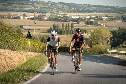 Zwei Radfahrer fahren auf einer Landstraße durch eine ländliche Gegend bei klarem Wetter
