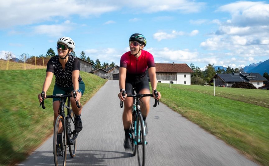 Due ciclisti pedalano su strada di campagna con case e montagne sullo sfondo