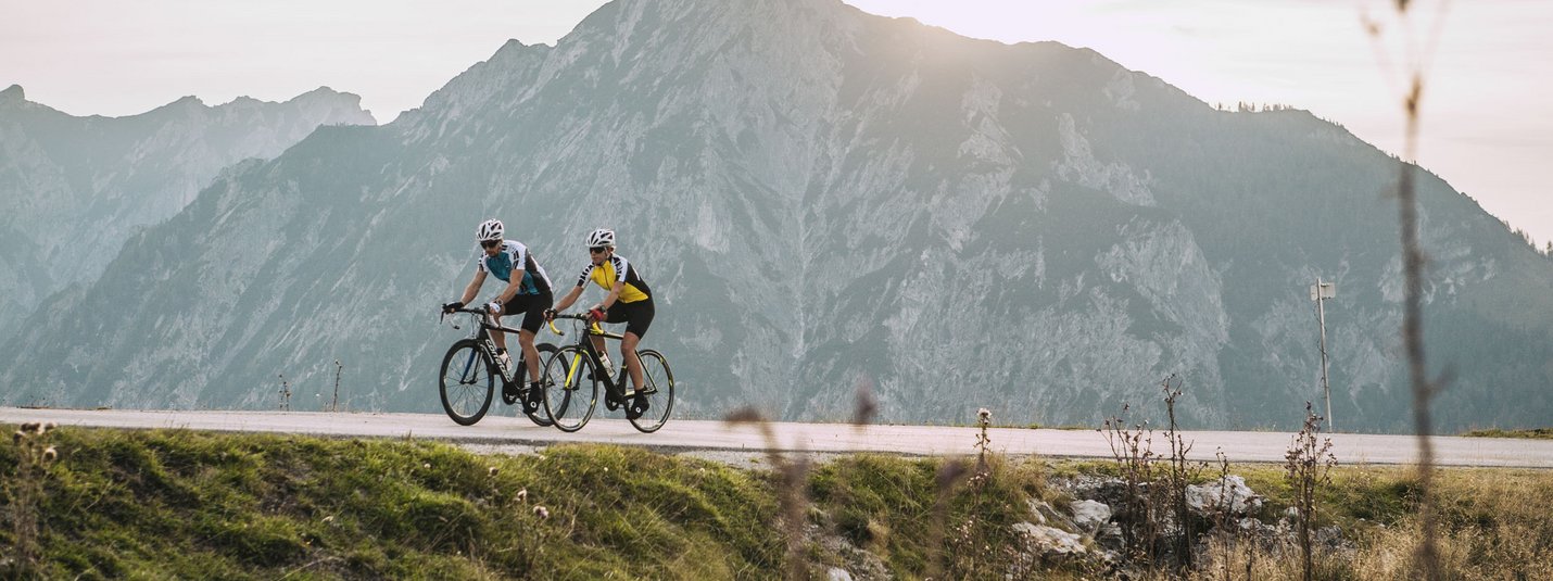 Two cyclists riding on mountain road with large mountain at sunset