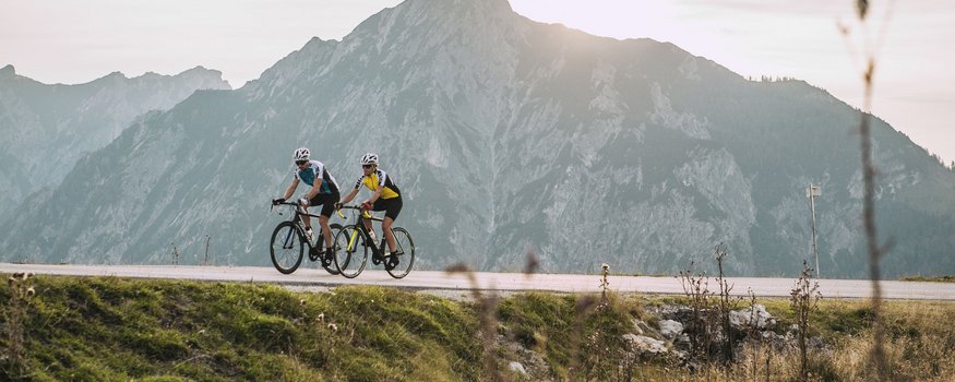 Zwei Radfahrer fahren auf Bergstraße mit großem Berg im Hintergrund bei Sonnenuntergang