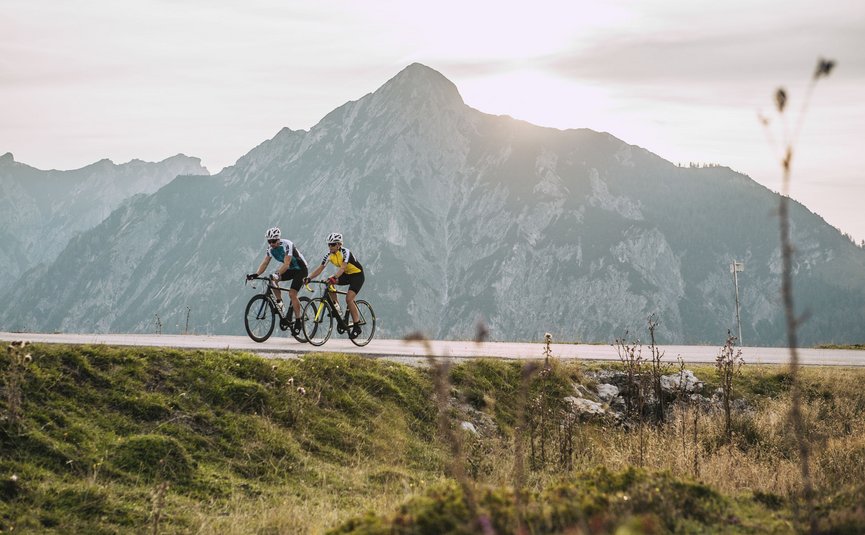 Two cyclists riding on mountain road with large mountain at sunset