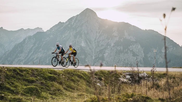 Die schönsten Rennrad-Regionen und Tipps für den Herbst © SalzburgerLand - Stefan Schopf for WOM Medien Zwei Radfahrer fahren auf Bergstraße mit großem Berg im Hintergrund bei Sonnenuntergang