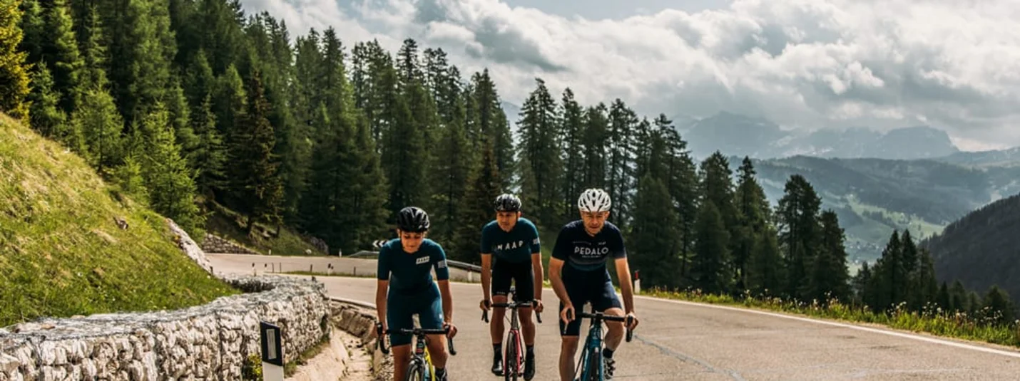 Road Bike Short Stay - 4 nights Tre ciclisti su strada di montagna con pini e cielo nuvoloso