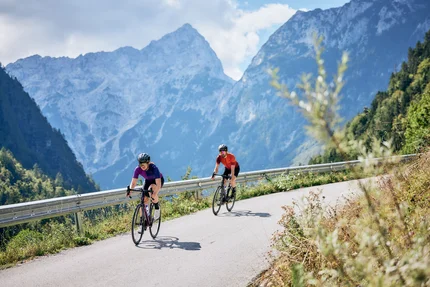 Zwei Radfahrer fahren auf bergiger Straße mit Alpen im Hintergrund