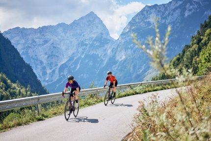 Zwei Radfahrer fahren auf bergiger Straße mit Alpen im Hintergrund