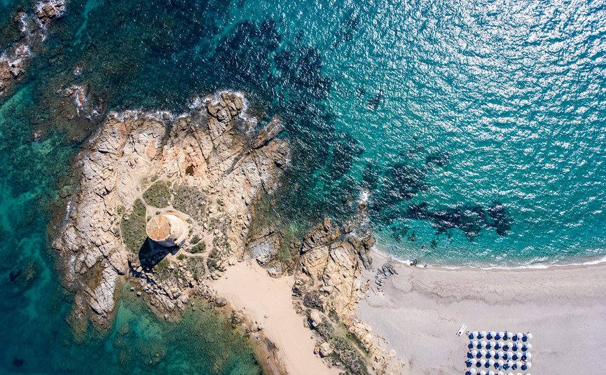 Aerial view of a rocky island with tower and beach umbrellas by the sea