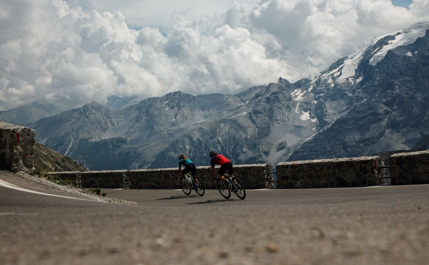 Two cyclists riding on a mountain road with snowy peaks behind them