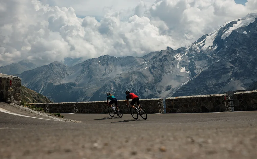 Due ciclisti su una strada di montagna con cime innevate sullo sfondo