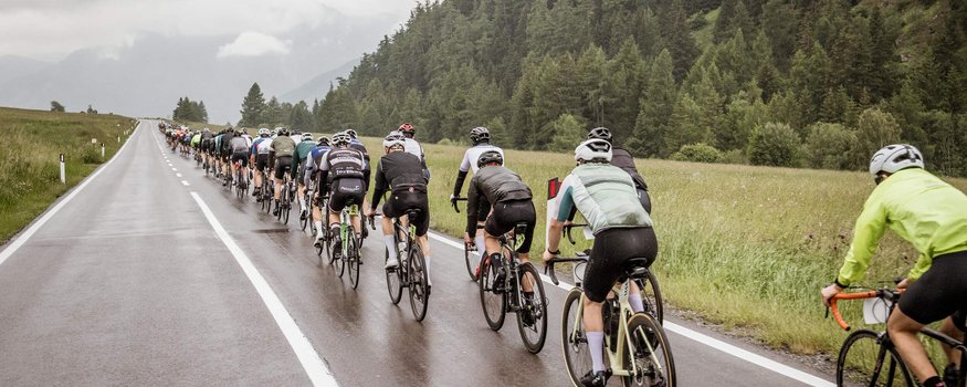 Dreiländergiro Nauders sul Passo di Resia © Fabian Schirgi Ciclisti su strada bagnata in campagna con foreste intorno