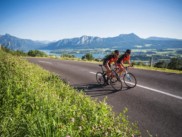 Ein Rennrad-Wochenende im SalzburgerLand – Salzkammergut © Heiko Mandl Zwei Radfahrer fahren auf einer Landstraße mit Bergen und See im Hintergrund