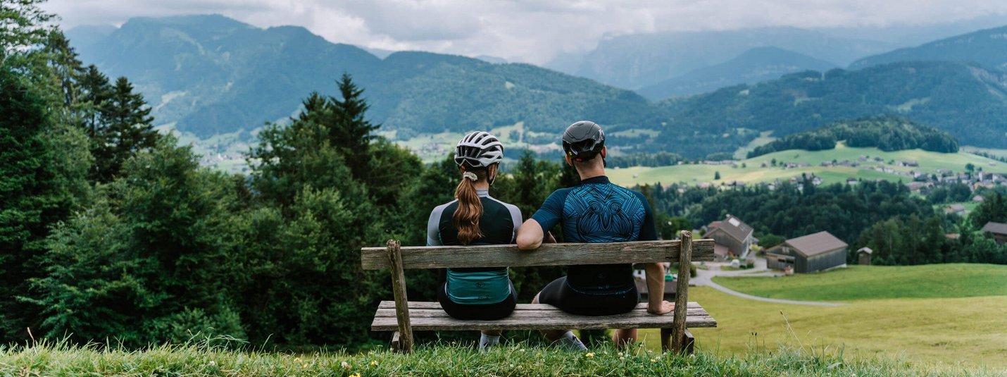 Twee fietsers op bank kijken uit over berglandschap met huizen en velden