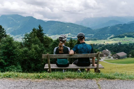 Zwei Radfahrer auf Bank blicken auf Berglandschaft mit Häusern und Wiesen