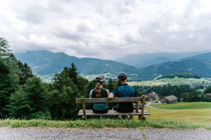 Zwei Radfahrer auf Bank blicken auf Berglandschaft mit Häusern und Wiesen