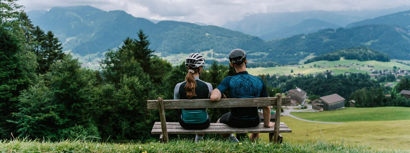Tour di familiarizzazione Bregenzerwald © Bregenzerwald Tourismus - Christina Neubauer Due ciclisti seduti su panchina guardano paesaggio montano con case e campi
