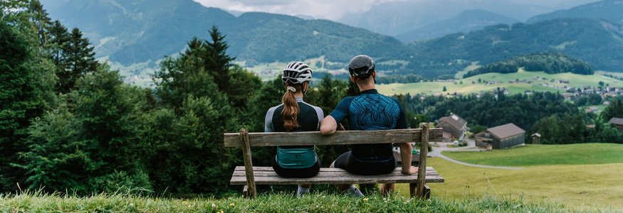 Zwei Radfahrer auf Bank blicken auf Berglandschaft mit Häusern und Wiesen