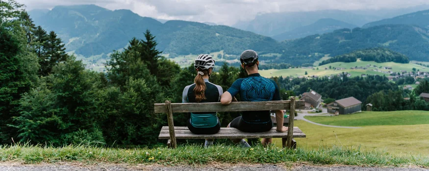 Zwei Radfahrer auf Bank blicken auf Berglandschaft mit Häusern und Wiesen