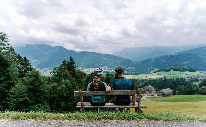 Due ciclisti seduti su panchina guardano paesaggio montano con case e campi