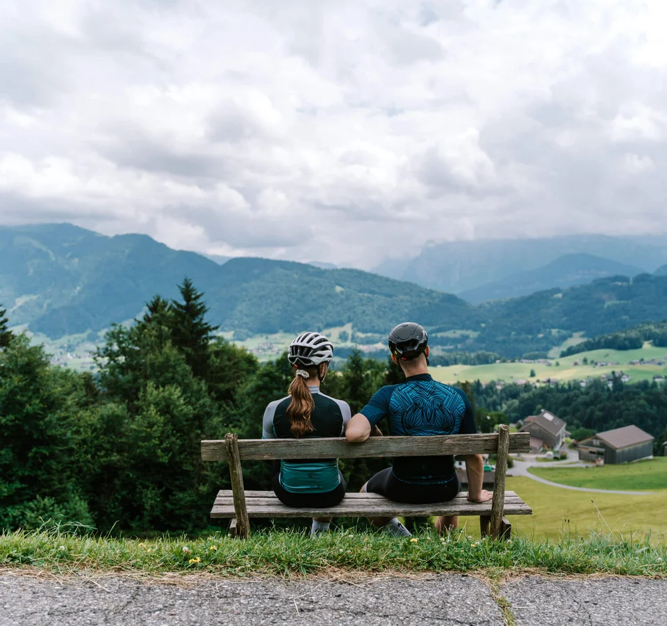A racing bike weekend in the Bregenzerwald © Bregenzerwald Tourismus - Christina Neubauer Two cyclists sitting on bench overlooking mountain landscape with houses and fields
