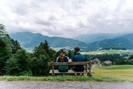 Zwei Radfahrer auf Bank blicken auf Berglandschaft mit Häusern und Wiesen