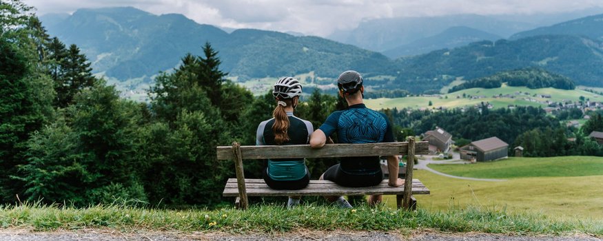 Zwei Radfahrer auf Bank blicken auf Berglandschaft mit Häusern und Wiesen