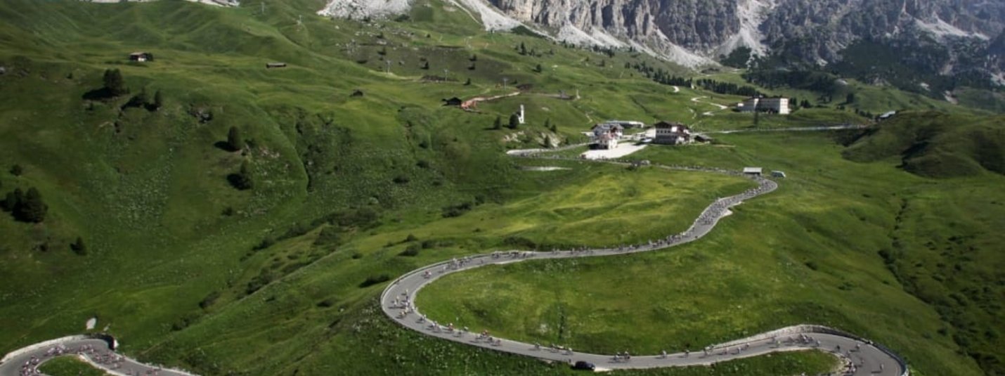 Sellaronda Bike Day - Juni Serpentinenstraße in grünen Bergen mit Radfahrern und Felsgipfeln im Hintergrund