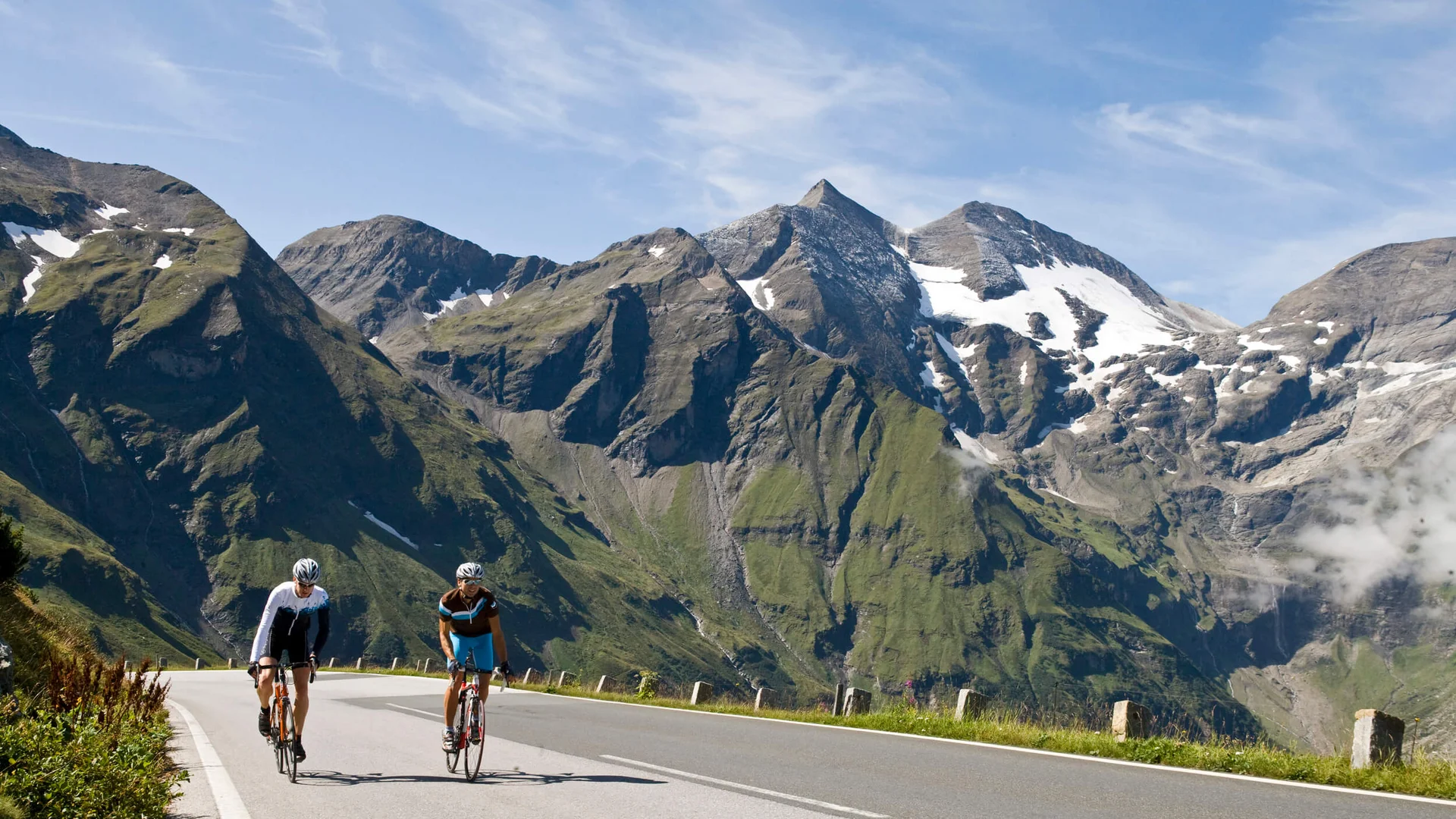 Zwei Radfahrer auf Bergstraße mit schneebedeckten Gipfeln im Hintergrund