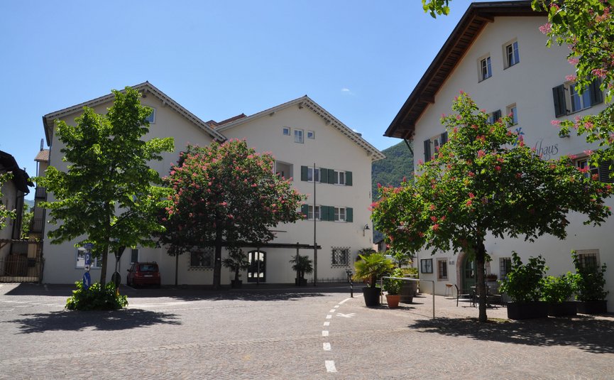 Sonniger Platz mit weißen Häusern, blühenden Bäumen und Bergen im Hintergrund
