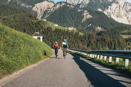 Twee fietsers rijden op bergweg met groene heuvels en rotsachtige bergen op de achtergrond