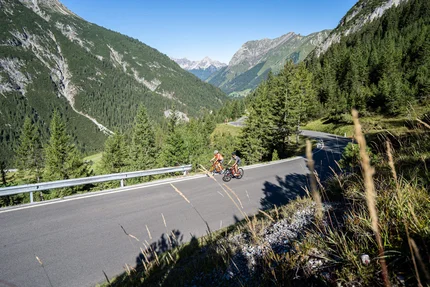 Zwei Radfahrer fahren auf Bergstraße in bewaldeter Alpenlandschaft bei klarem Himmel