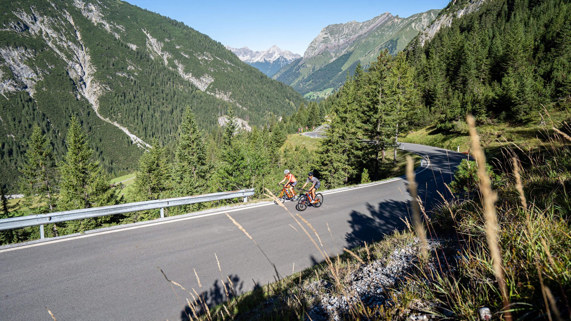 Two cyclists riding on a mountain road in a forested alpine landscape on a clear day
