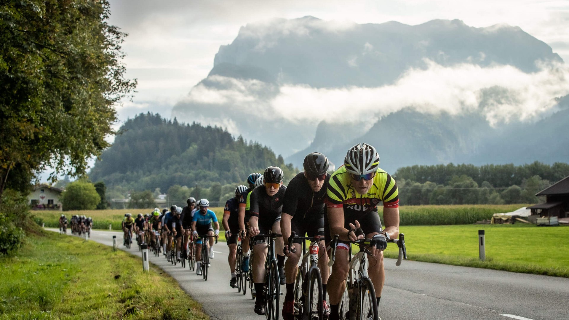 Radfahrer fahren in einer Gruppe auf einer Landstraße vor Bergen und Wolken
