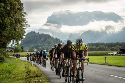 Radfahrer fahren in einer Gruppe auf einer Landstraße vor Bergen und Wolken