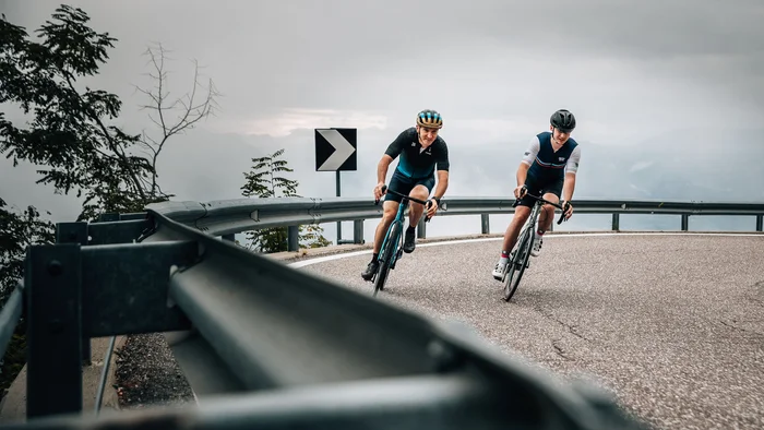 Zwei Radfahrer fahren eine kurvige Bergstraße bei bewölktem Himmel hinab