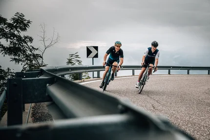Zwei Radfahrer fahren eine kurvige Bergstraße bei bewölktem Himmel hinab