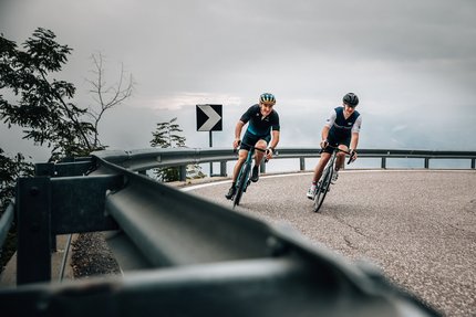 Twee wielrenners dalen een kronkelende bergweg af bij bewolkte lucht