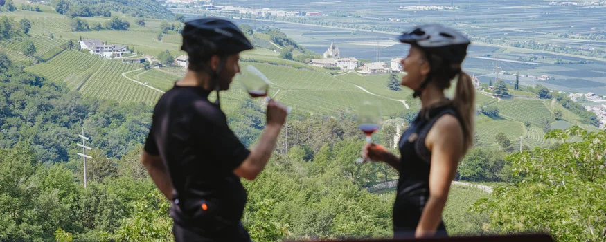 Saisonstart im Teutschhaus © Thomas Monsorno - TV Castelfeder Zwei Radfahrer trinken Wein mit Blick auf grüne Weinberge und Berge