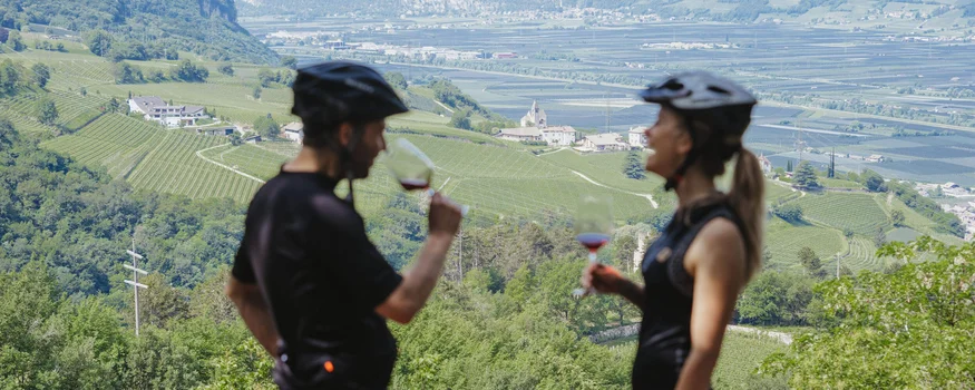 Kurven, Kultur & Kontraste in Südtirols Süden © Thomas Monsorno - TV Castelfeder Zwei Radfahrer trinken Wein mit Blick auf grüne Weinberge und Berge