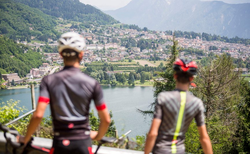 Due ciclisti osservano un lago con città e montagne sullo sfondo