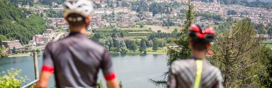 Due ciclisti osservano un lago con città e montagne sullo sfondo