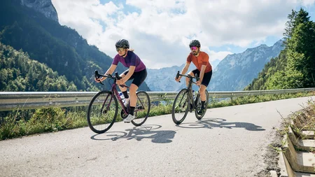 Due ciclisti pedalano su strada di montagna con cielo sereno e nuvole