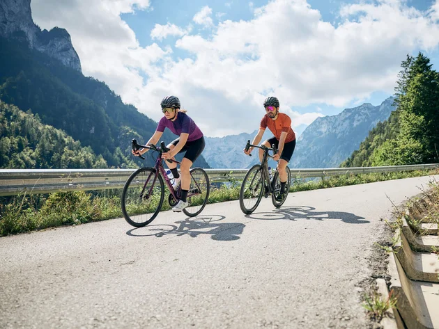 Due ciclisti pedalano su strada di montagna con cielo sereno e nuvole